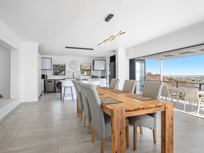A modern kitchen with a central island, a dining area with a wooden table and grey chairs, and a large window offering a view of the landscape.