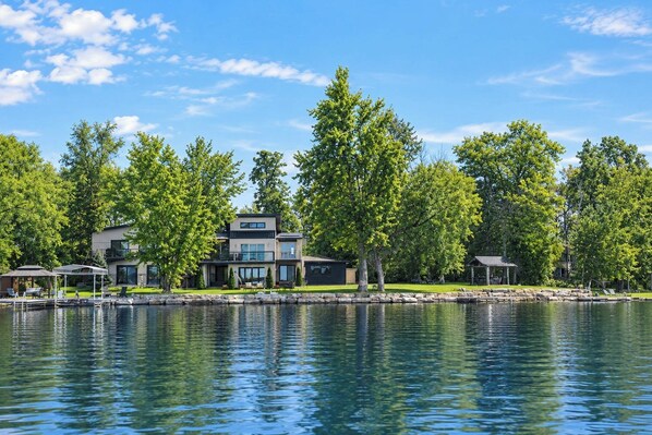 A modern two-story house with a dock extending into a calm lake, surrounded by lush green trees.