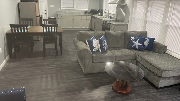 Een moderne woonkamer met een modulaire bank, een eettafel met stoelen, een keuken met fornuis en spoelbak, en een groot raam met jaloezieën.
