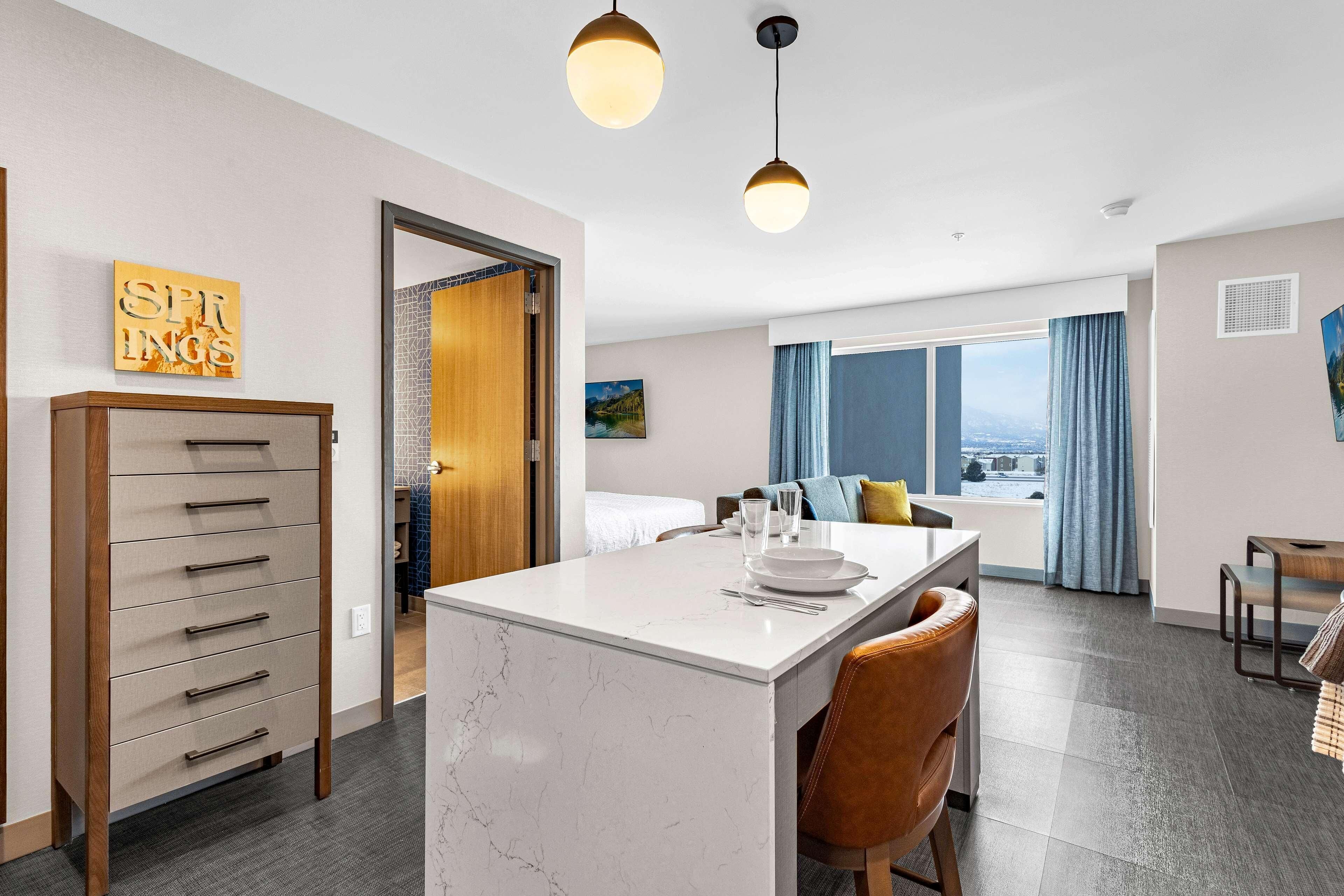 Una habitación de hotel moderna con una isla de mármol, un aparador de madera, un comedor y un ventanal con vistas.