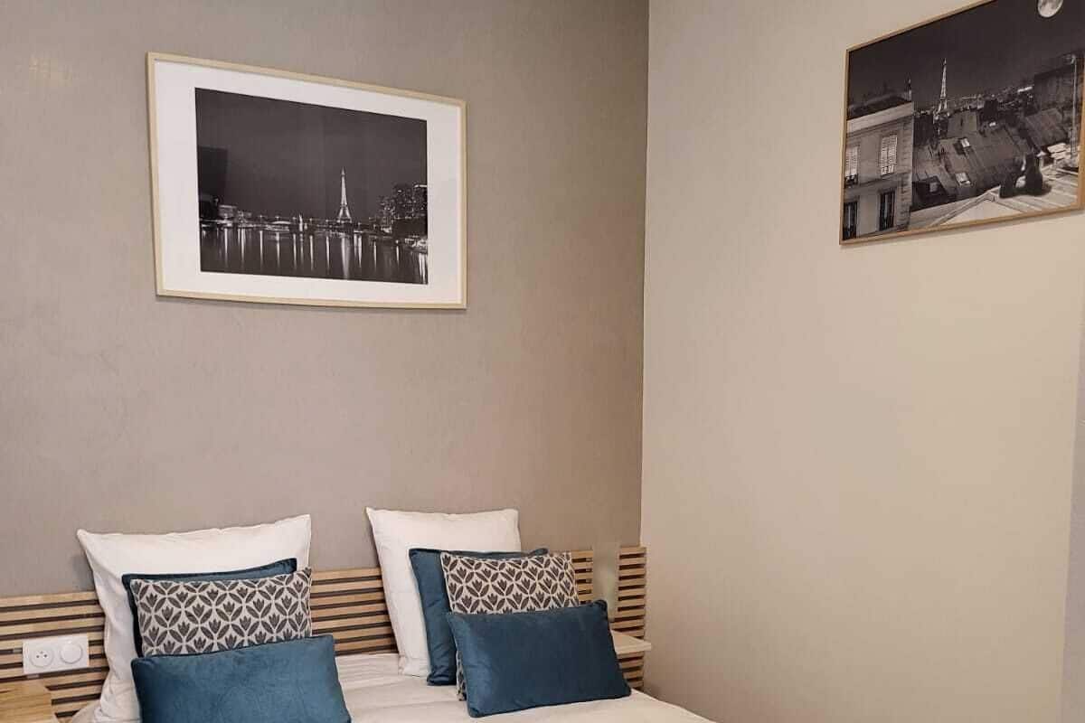 Un lit bien fait, recouvert d’une couvre-lit turquoise et agrémenté de deux oreillers, avec un tableau représentant un paysage urbain encadré au-dessus, et une table de chevet avec une lampe.