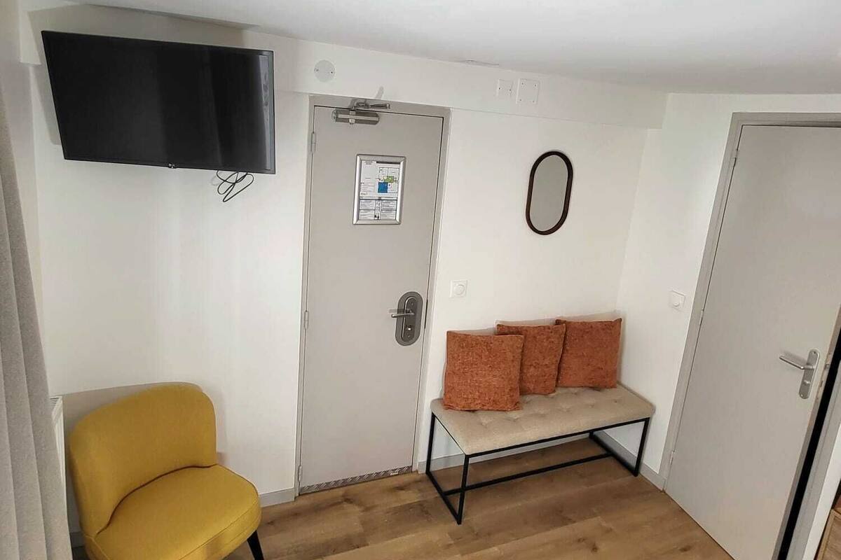 Une petite pièce meublée, avec une chaise jaune, un banc avec des coussins, une table ronde en bois et un téléviseur à écran plat fixé au mur.