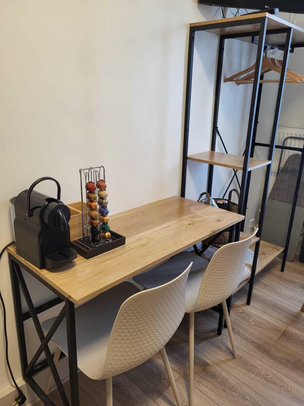 Un pequeño comedor con una mesa de madera, dos sillas blancas, una máquina de café negra y un estante metálico con varios objetos.