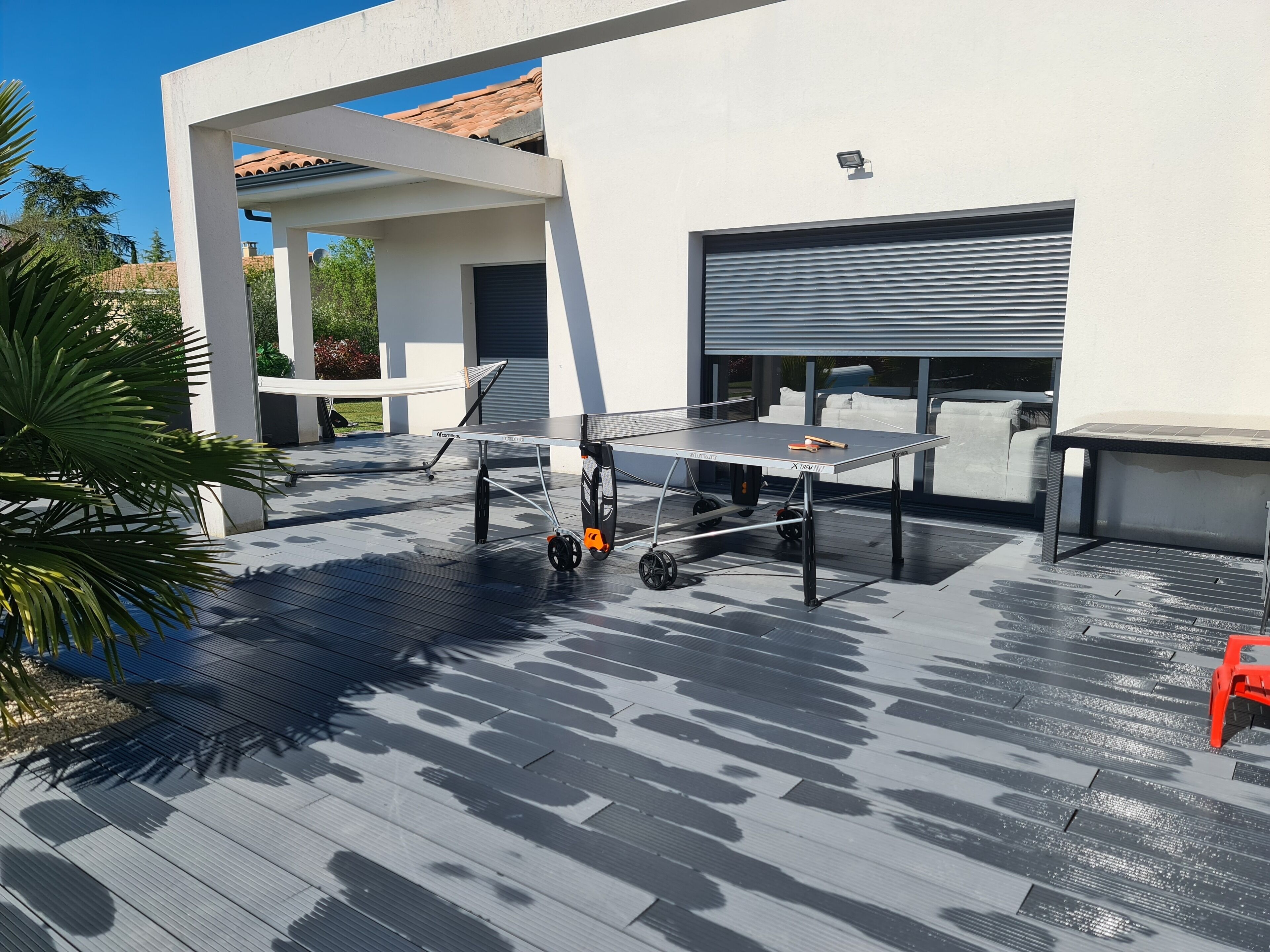 An outdoor table tennis table with wheels, set up on a wooden deck with a modern house in the background.