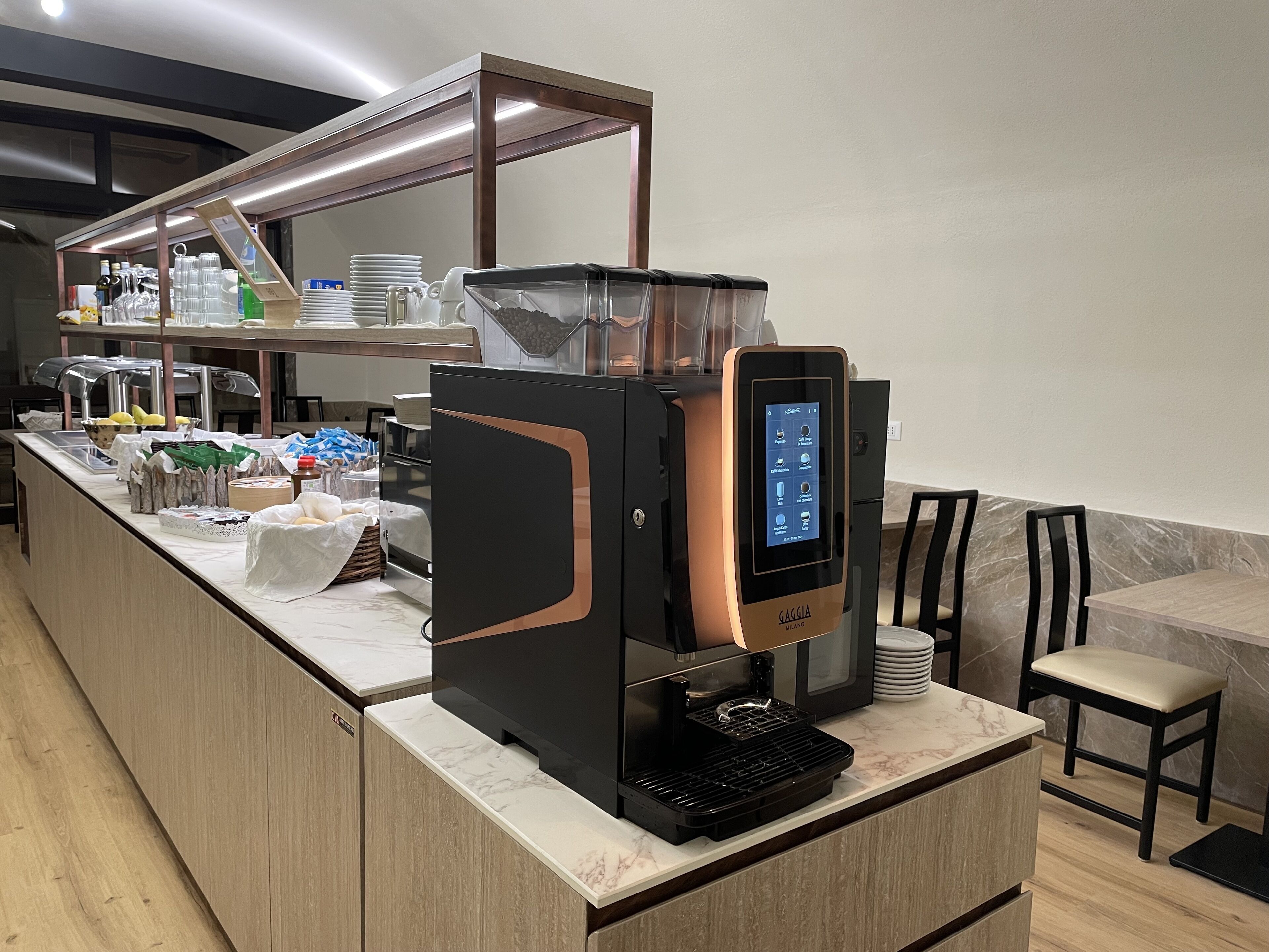 A self-service coffee station with a touchscreen interface, a coffee machine, and a variety of coffee pods and milk options.