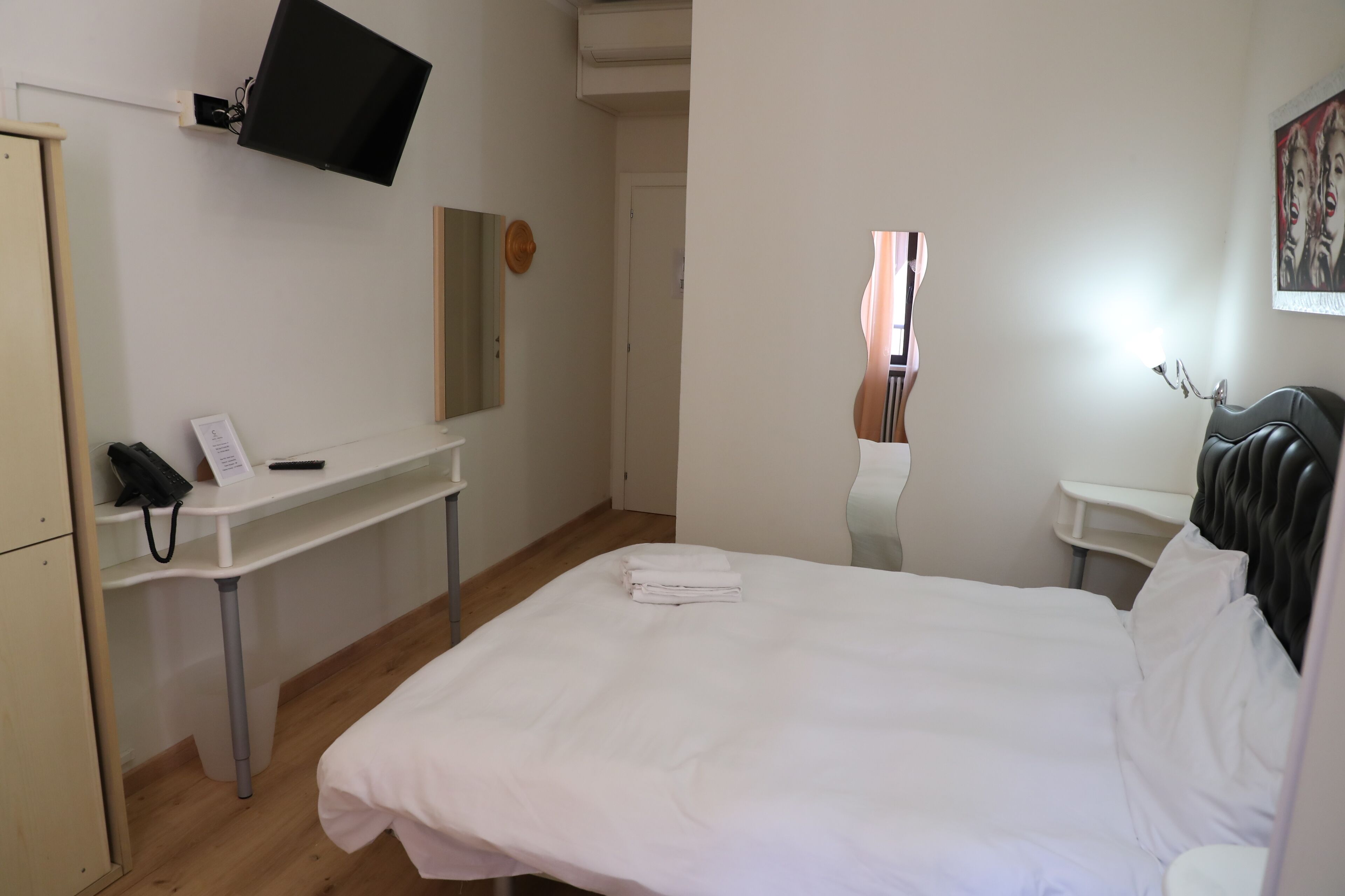 A hotel room with a bed, a desk with a telephone, a flat-screen TV mounted on the wall, and a mirror.