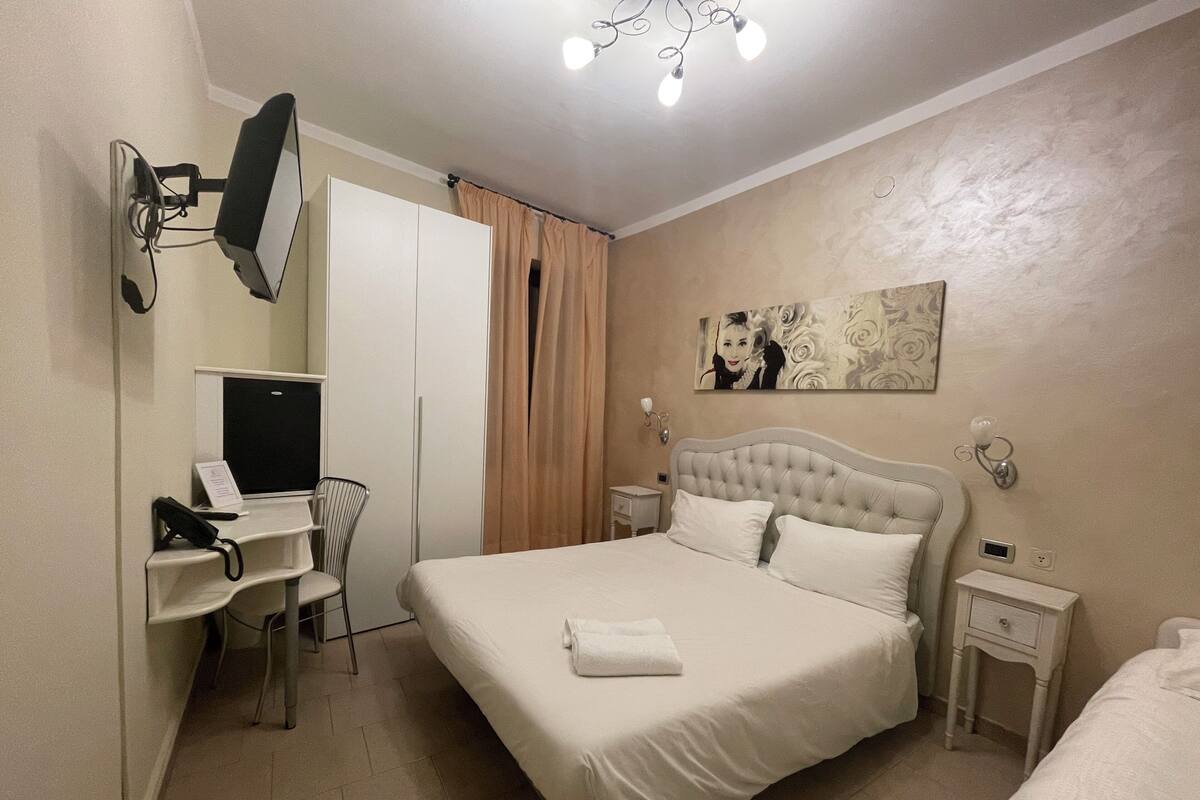 A hotel room with a bed, a desk with a computer, a chair, a wardrobe, and a painting on the wall.