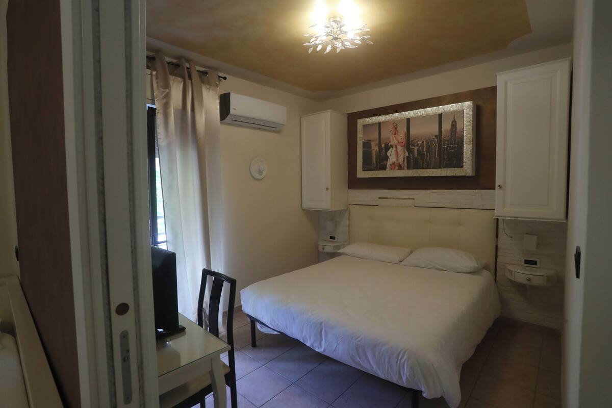 A bedroom with a bed, a desk, a chair, an air conditioner, a nightstand, a wall clock, and a painting.