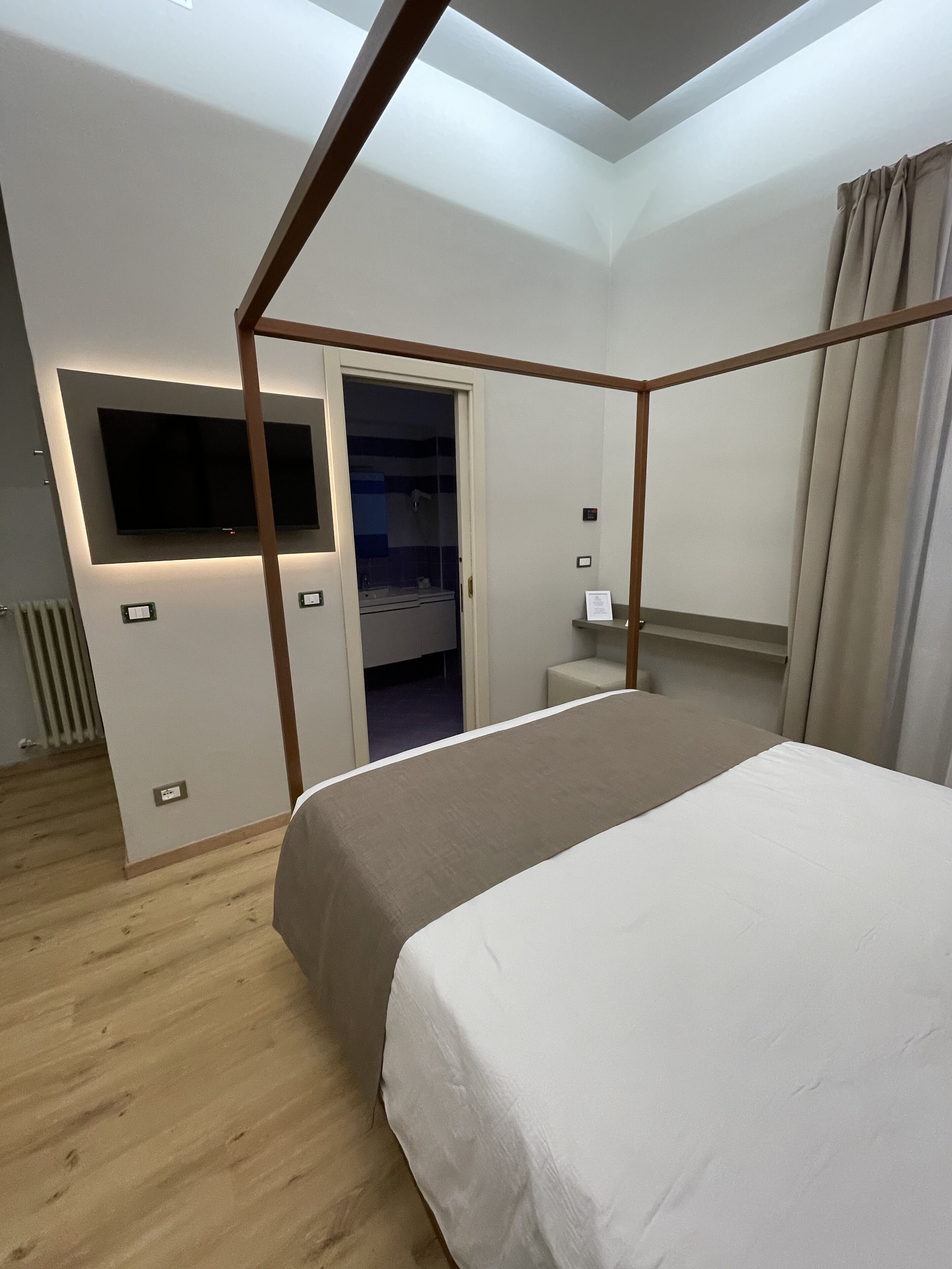 Et moderne hotelværelse med himmelseng, et fladskærms-tv monteret på væggen og et separat badeværelse, der kan anes gennem en åben dør.