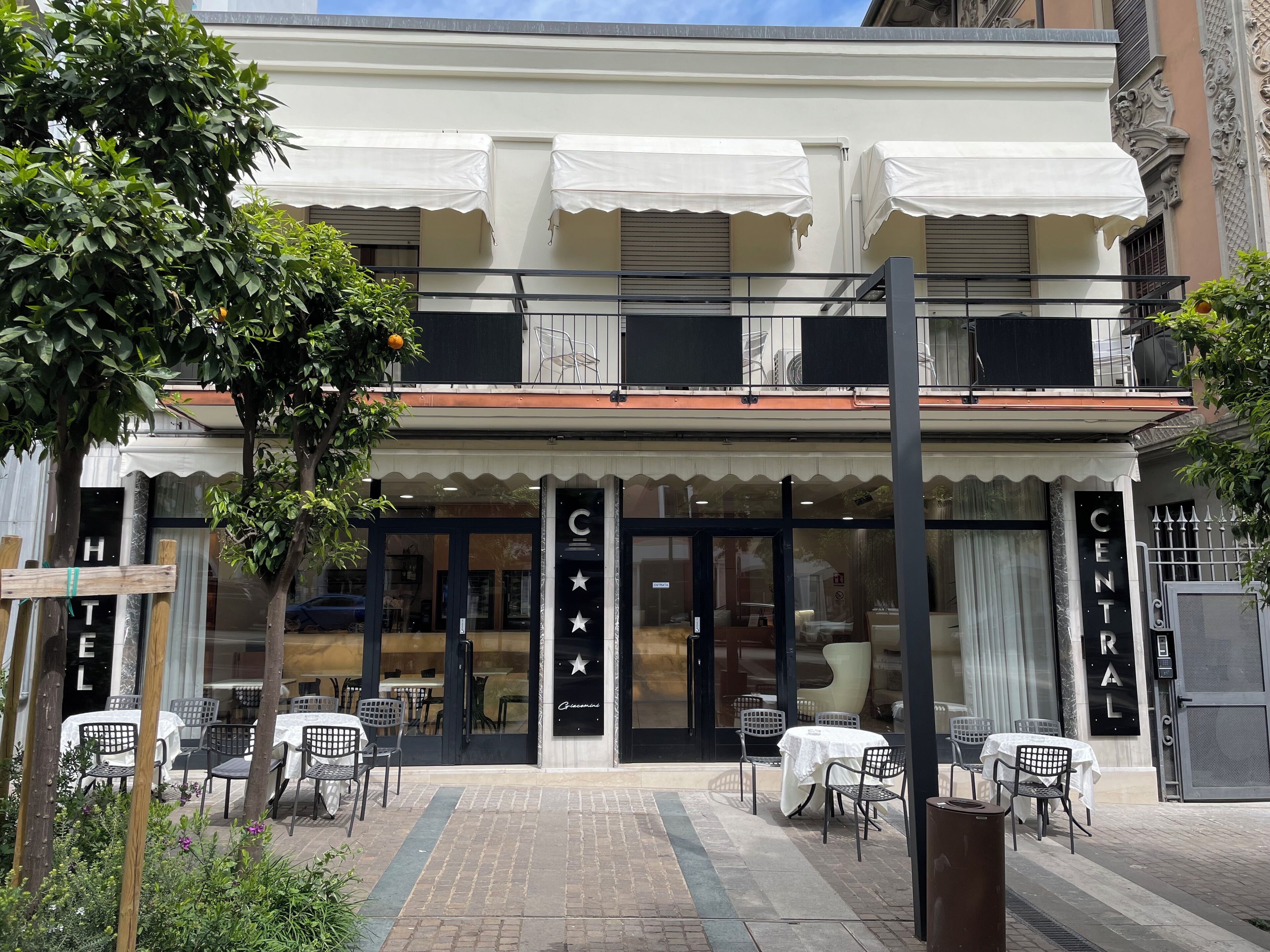 A hotel with a central entrance, outdoor seating, and a balcony with white awnings.