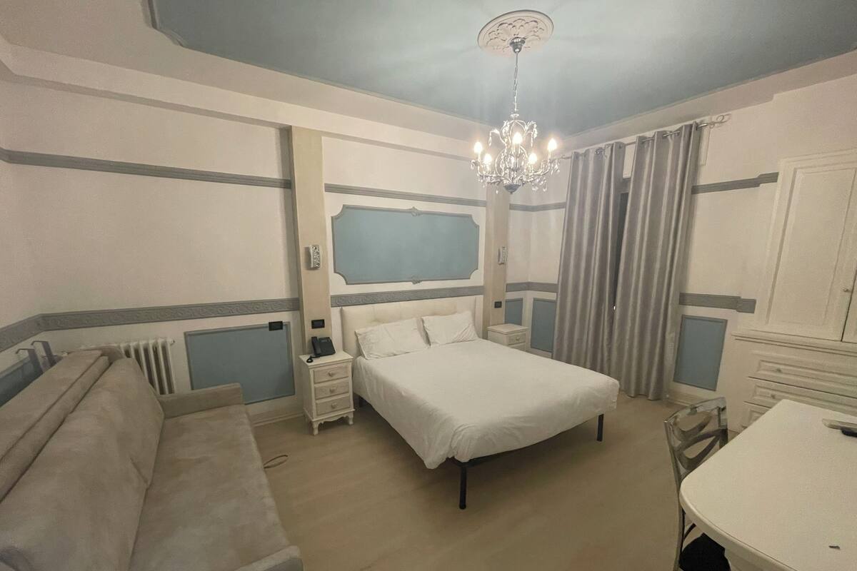 A hotel room with a bed, bedside tables, a desk, and a chandelier.