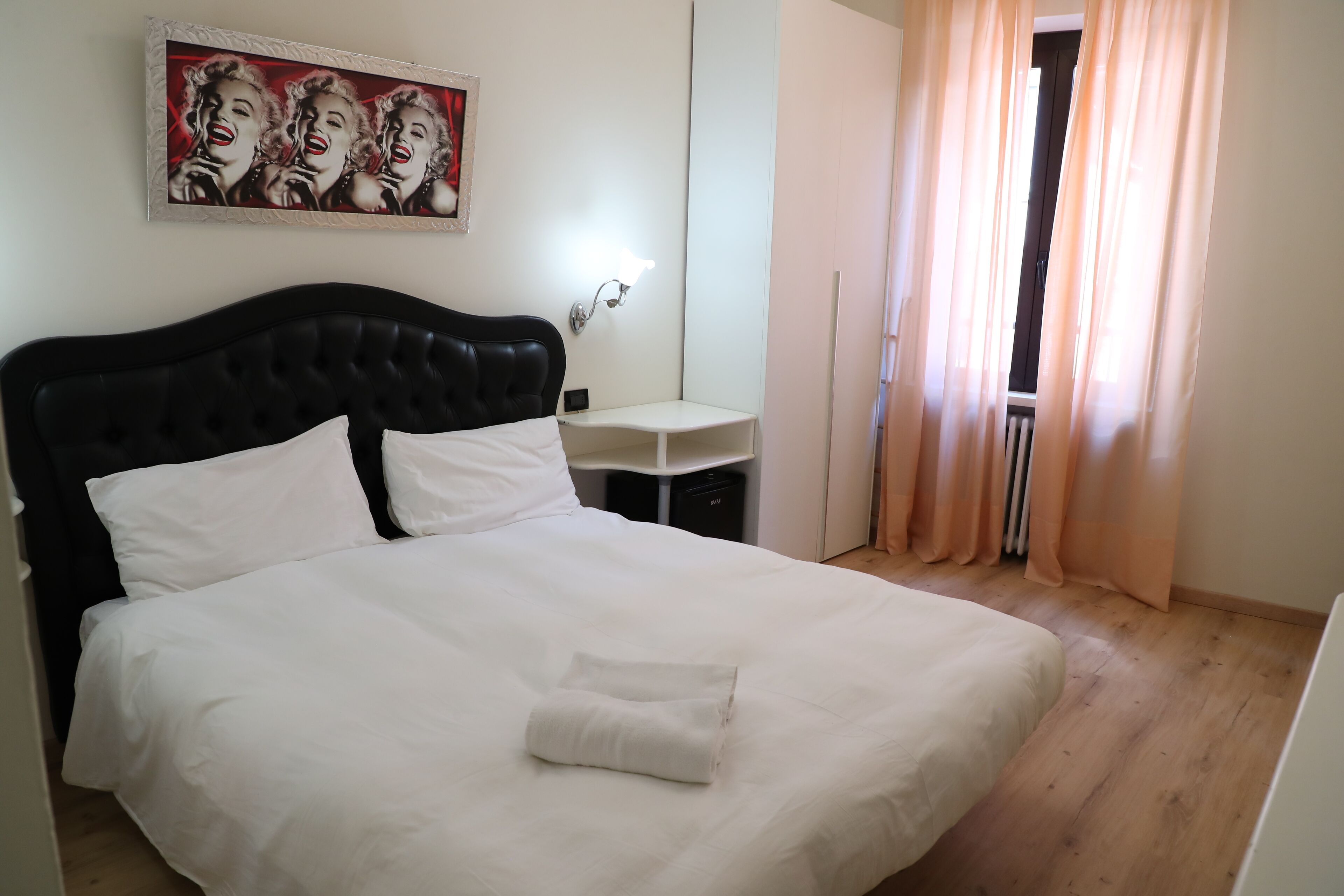 Un lit bien fait, avec du linge de lit blanc, une tête de lit noire, une petite table de chevet avec une lampe, et une œuvre d’art encadrée au-dessus du lit.