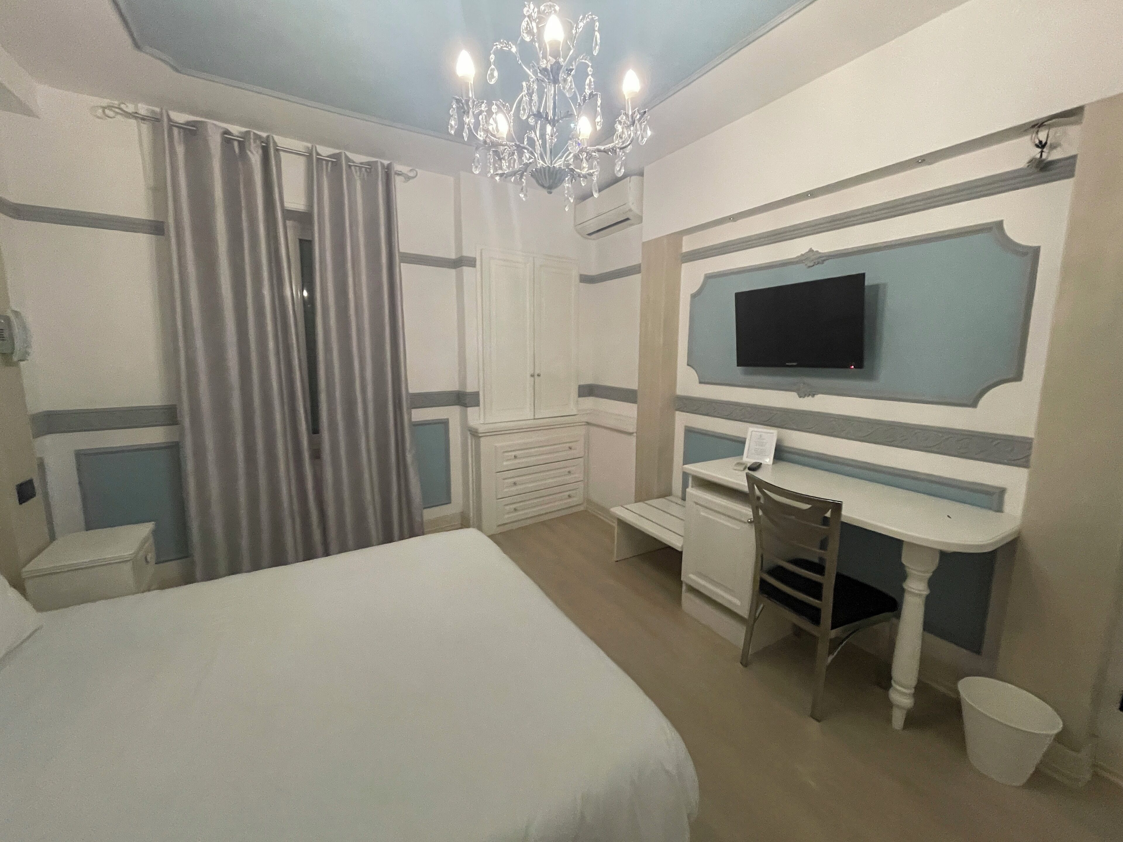 A hotel room with a bed, a desk, a chair, a television, and a chandelier.