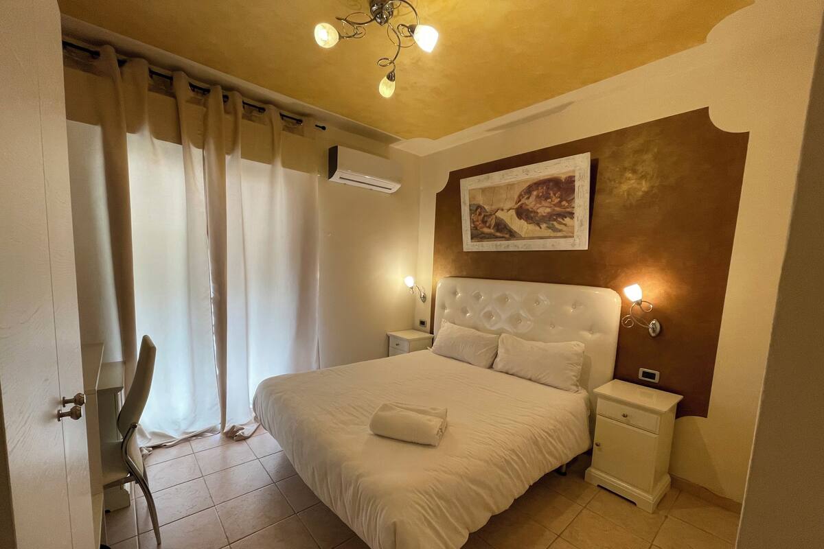 A bedroom with a large bed, a nightstand, a wall-mounted air conditioner, and a painting above the bed.