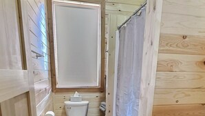 Une petite salle de bain étroite, avec un plancher en bois, des toilettes, une fenêtre et un rideau de douche.