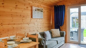A wooden cabin interior with a dining table set for two, a sofa, and a sliding glass door leading to an outdoor area.