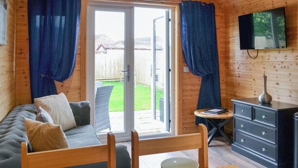 A cozy room with a sofa, dining table set with plates and cutlery, a television mounted on the wall, and a view of a garden through the open door.