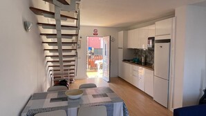 A compact kitchen with white cabinets, a refrigerator, and a microwave. There is a dining table with chairs and a patterned tablecloth. A staircase with wooden steps leads to an open door.