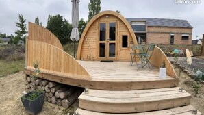Une terrasse en bois dotée d’une structure de toit incurvée, un petit espace salon avec des chaises et une porte donnant accès à un bâtiment.