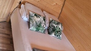 Une chambre chaleureuse avec un lit, deux oreillers ornés d’un motif de feuilles tropicales, un oreiller matelassé gris et des poutres en bois au plafond.