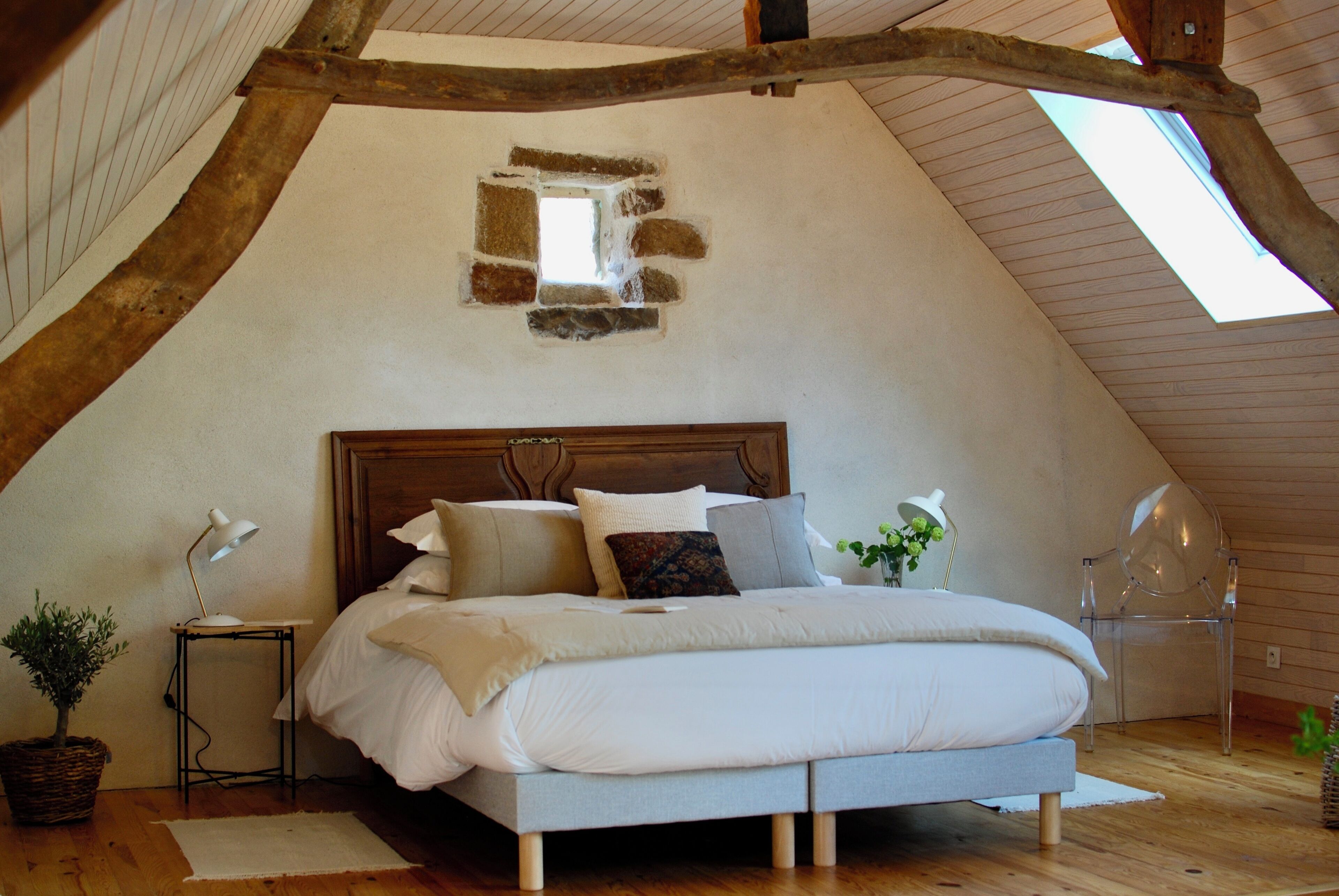 A bedroom with a sloped ceiling, a wooden headboard, a bed with white bedding, a small table with a lamp, and a potted plant.