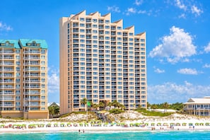 Un hotel frente a la playa con varios pisos, balcones y una zona de arena con sombrillas.