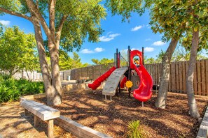 Un parque infantil con tobogán y estructuras para escalar, rodeado de árboles y una valla de madera.