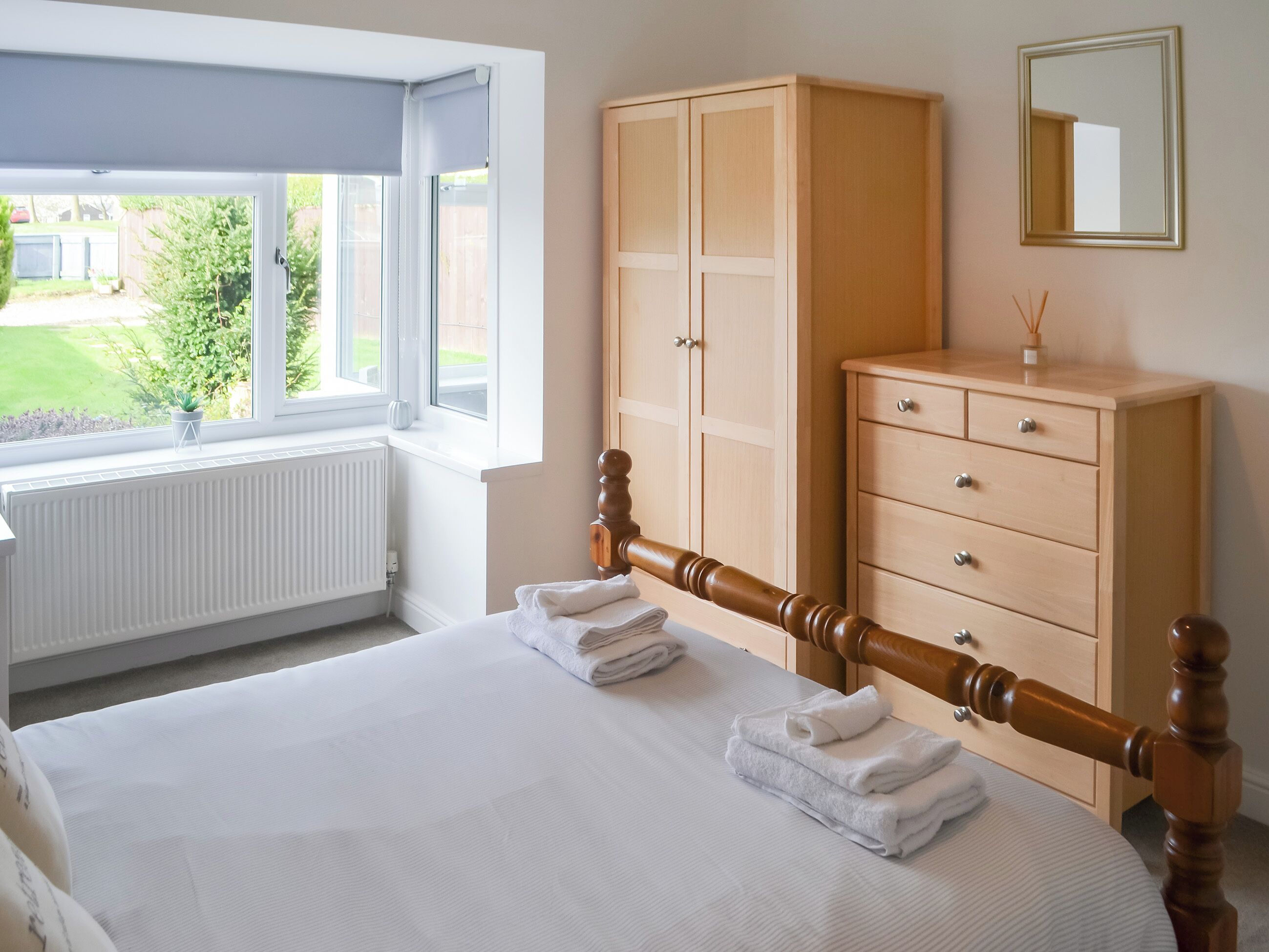 A bedroom with a bed, a wooden wardrobe, a radiator, and a window with a view of greenery.
