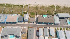 Vista aérea de un hotel frente a la playa con varias construcciones, una carretera y una playa con sombrillas.
