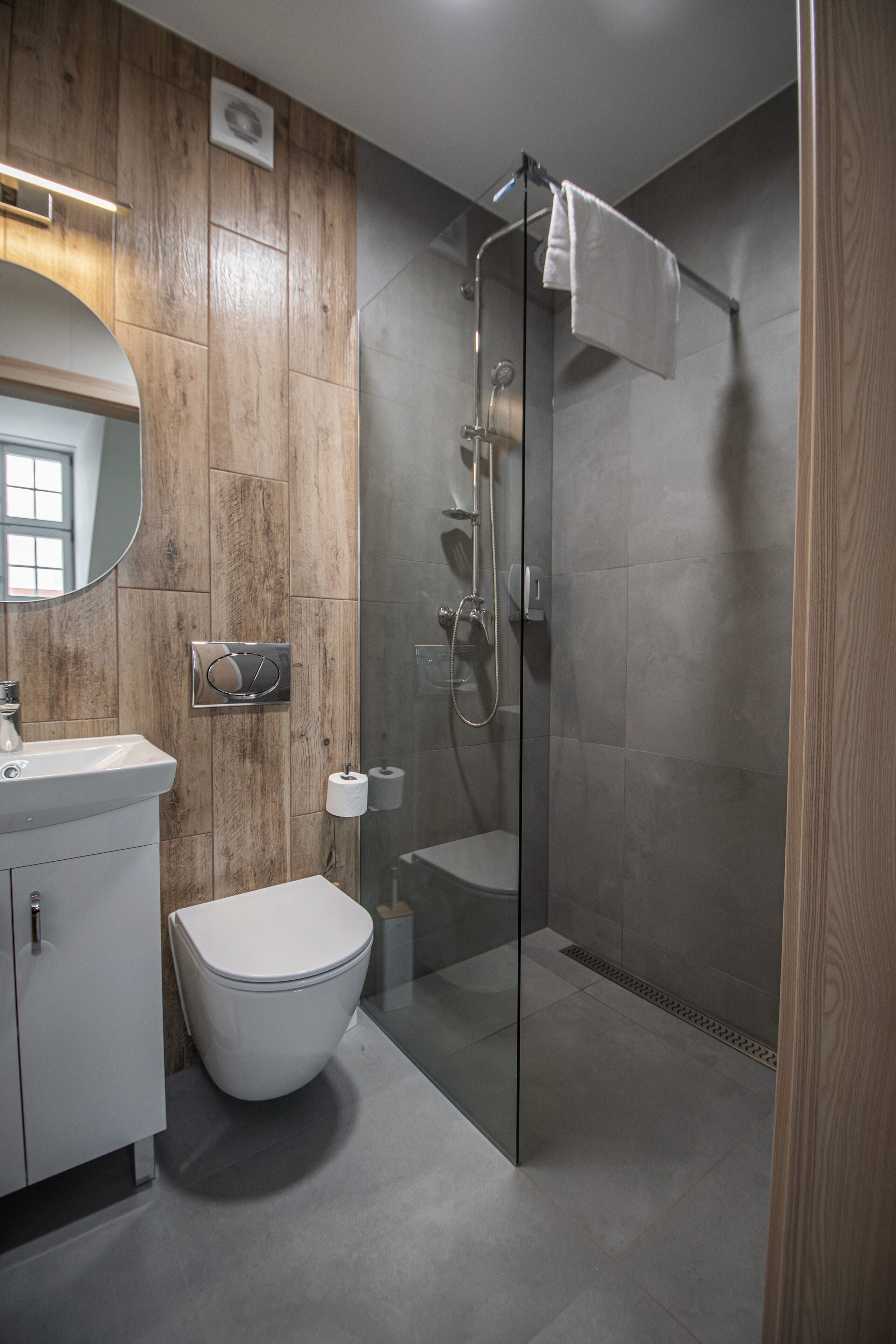 Une salle de bain moderne avec un mur recouvert de panneaux en bois, des toilettes blanches, une douche en verre et un lavabo blanc.