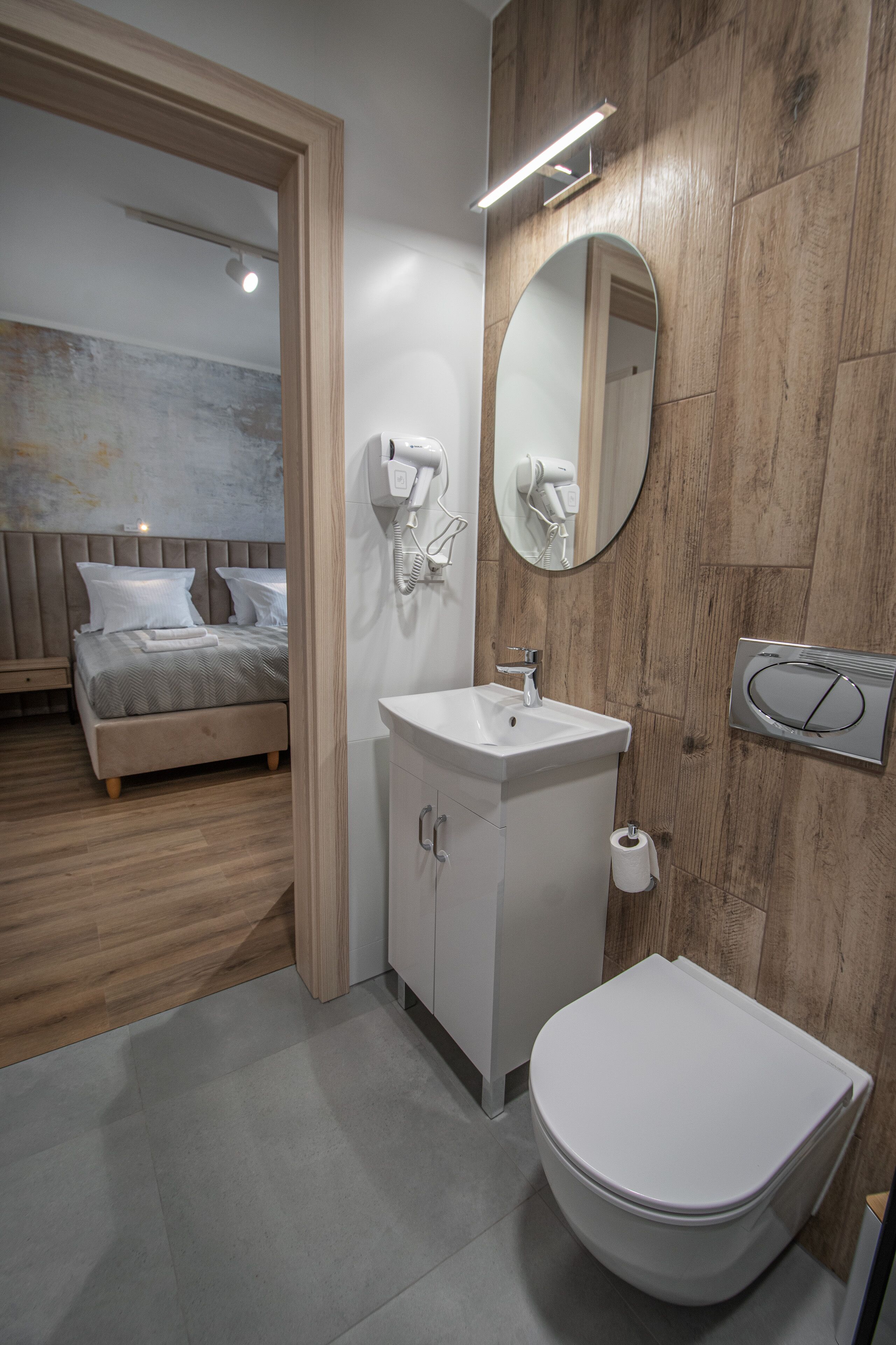 A modern bathroom with a wooden wall, a white sink, a toilet, and a bed visible through an open door.