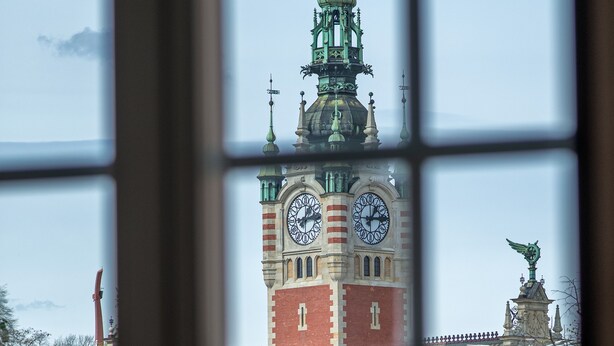 Une vue à travers la fenêtre d’un bâtiment doté d’une tour horlogée, d’une statue et donnant sur une rue bordée d’arbres et où circulent des voitures.