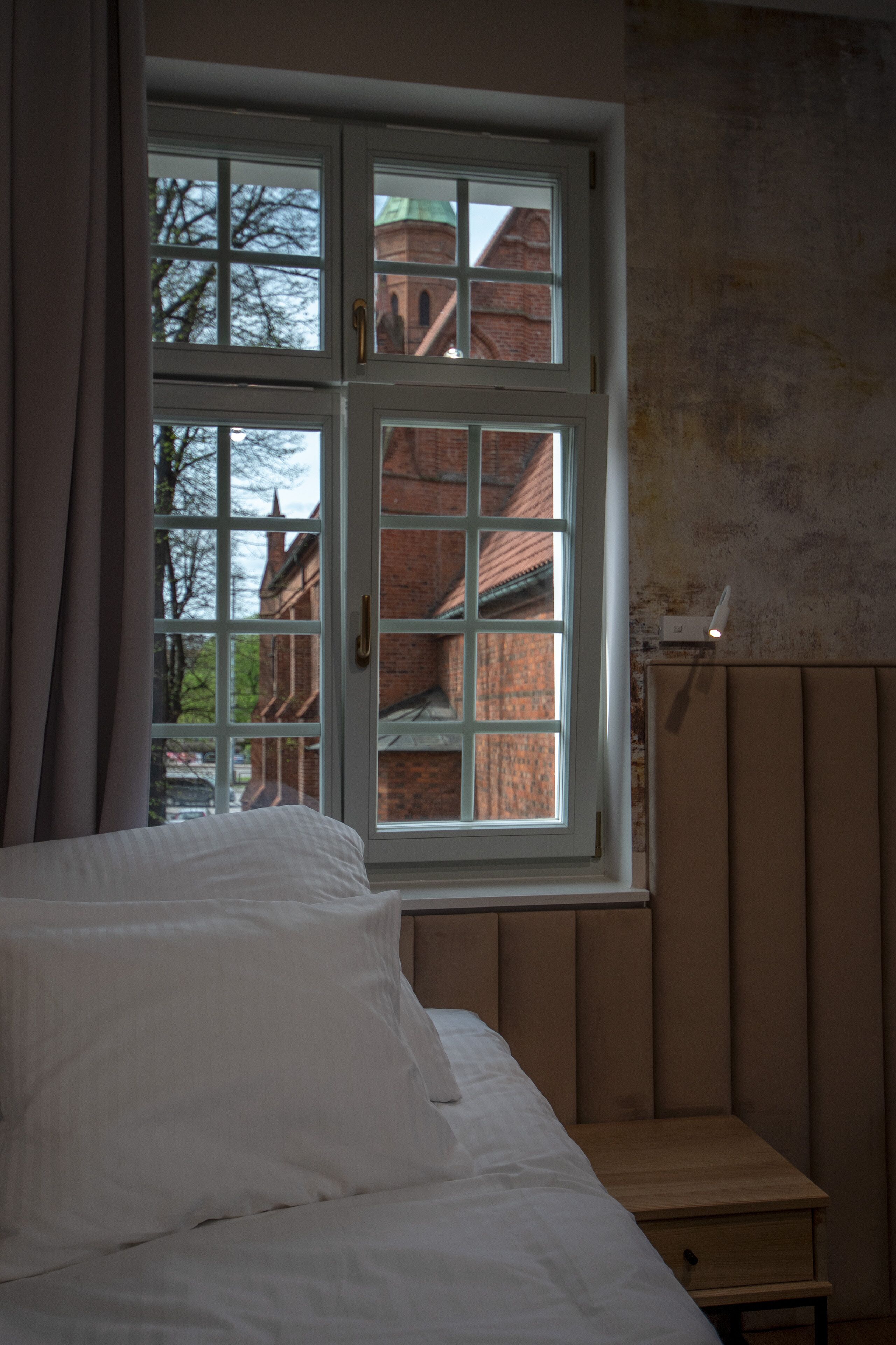 A bedroom with a large window, a bed, a bedside table, and a wall-mounted light fixture.