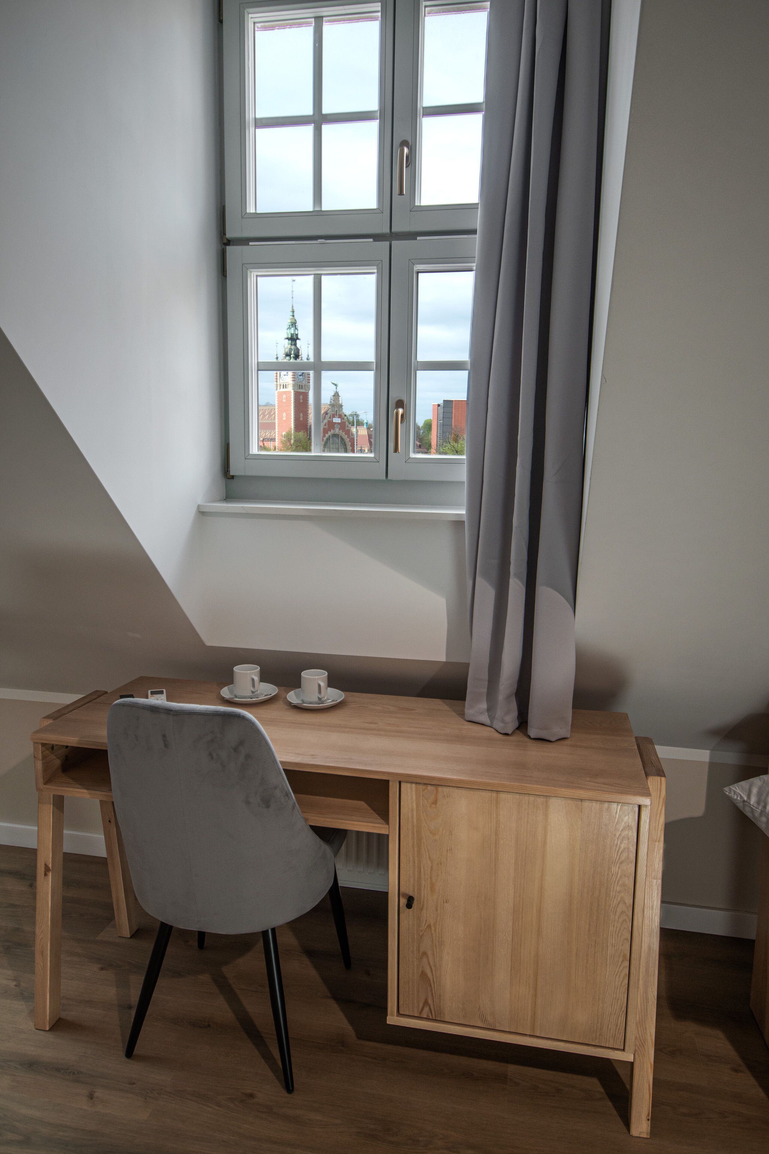 A room with a wooden desk, two white cups, a grey chair, and a window with curtains.