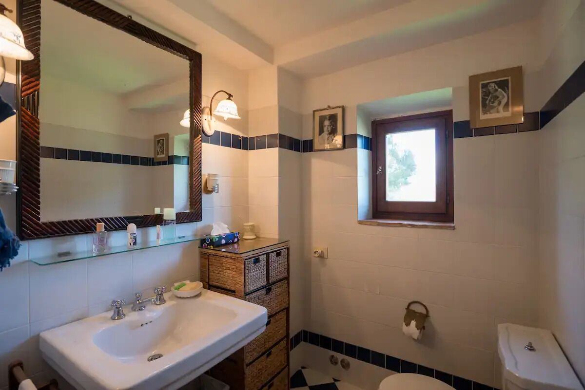 A bathroom with a white sink, a mirror, and a toilet. There are two framed pictures on the wall, and a window above the toilet.