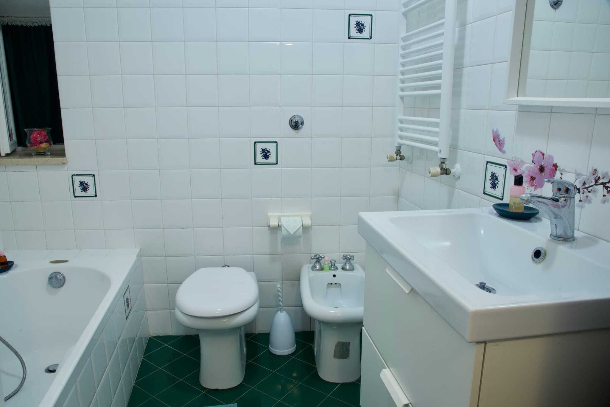 Et badeværelse med hvide fliser, grøn gulv, et badekar, et toilet, en bidet og en vask med et spejl.