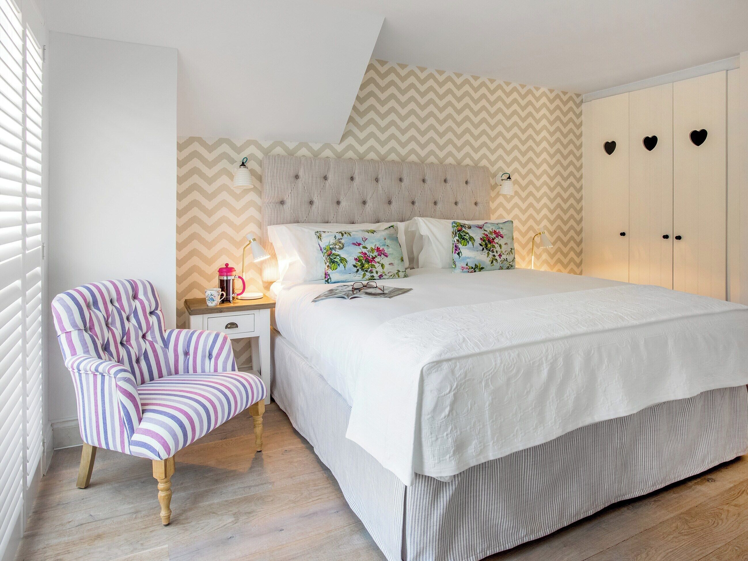 A bedroom with a bed, a striped armchair, a bedside table with a lamp, and a wardrobe with heart-shaped cutouts.