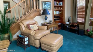 Un salon avec un fauteuil en cuir, un escalier en bois, une bibliothèque et une fenêtre avec des stores.