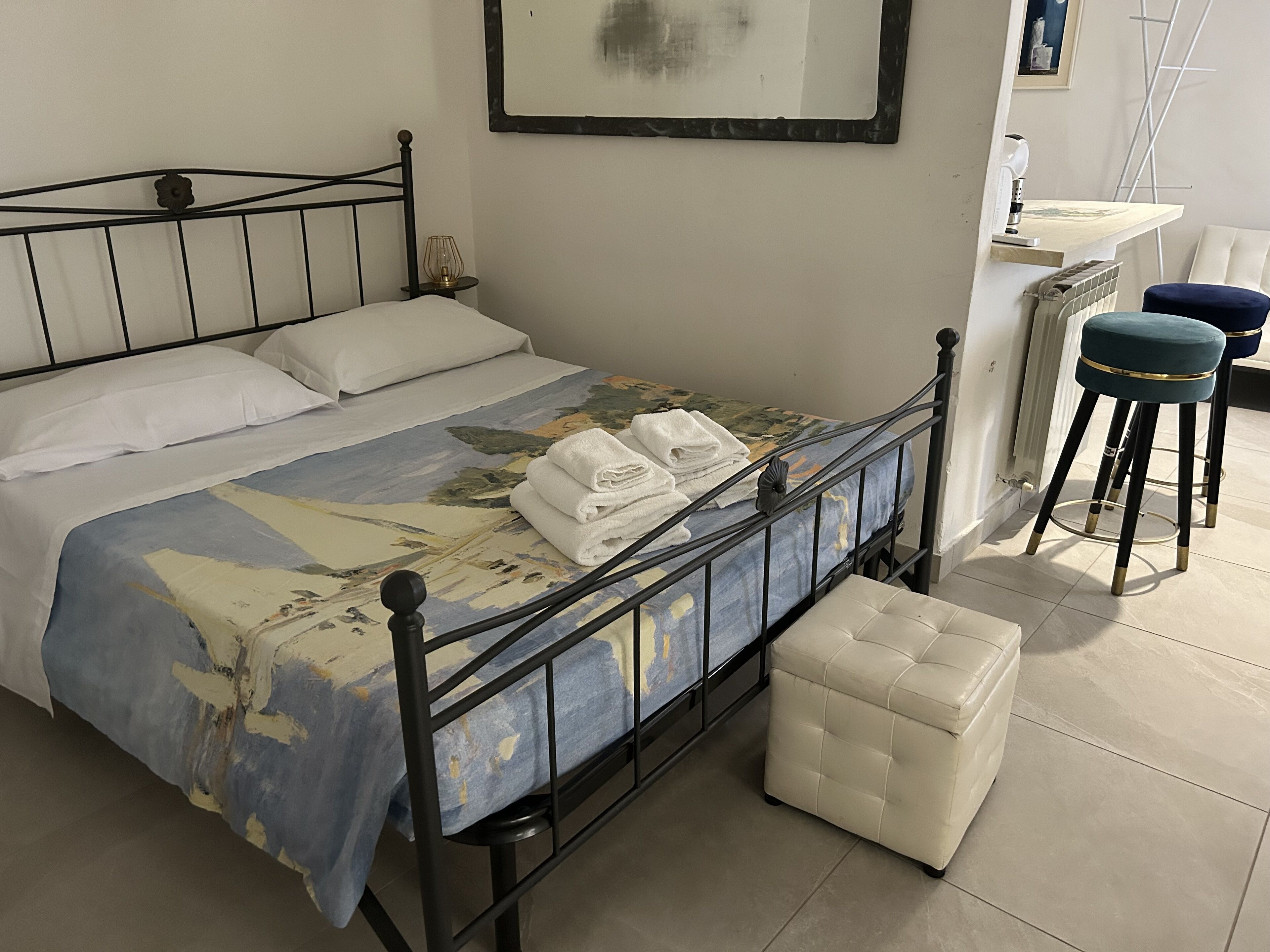 A neatly made bed with a patterned bedspread, a bedside table with a lamp, a small stool, and a chair.