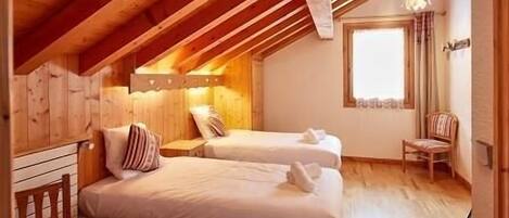 A cozy attic bedroom with two beds, wooden beams, a window, and a chair.