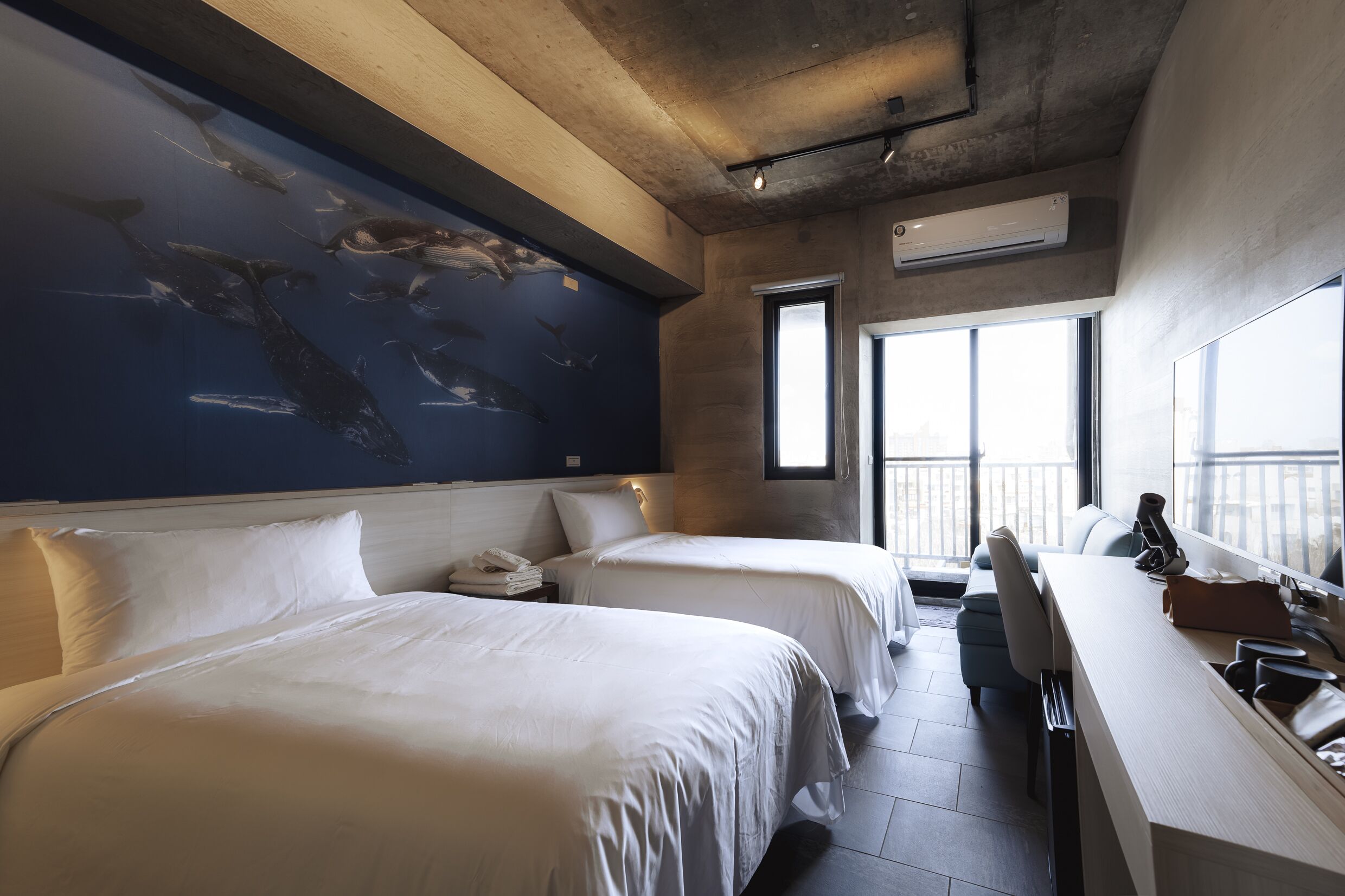 A hotel room with two beds, a desk, a chair, a large window, and a mural of whales on the wall.