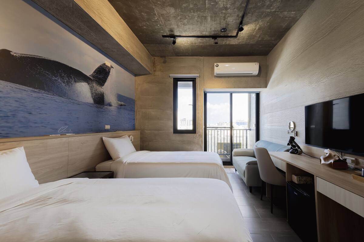 A hotel room with two beds, a desk, a TV, and a large window.