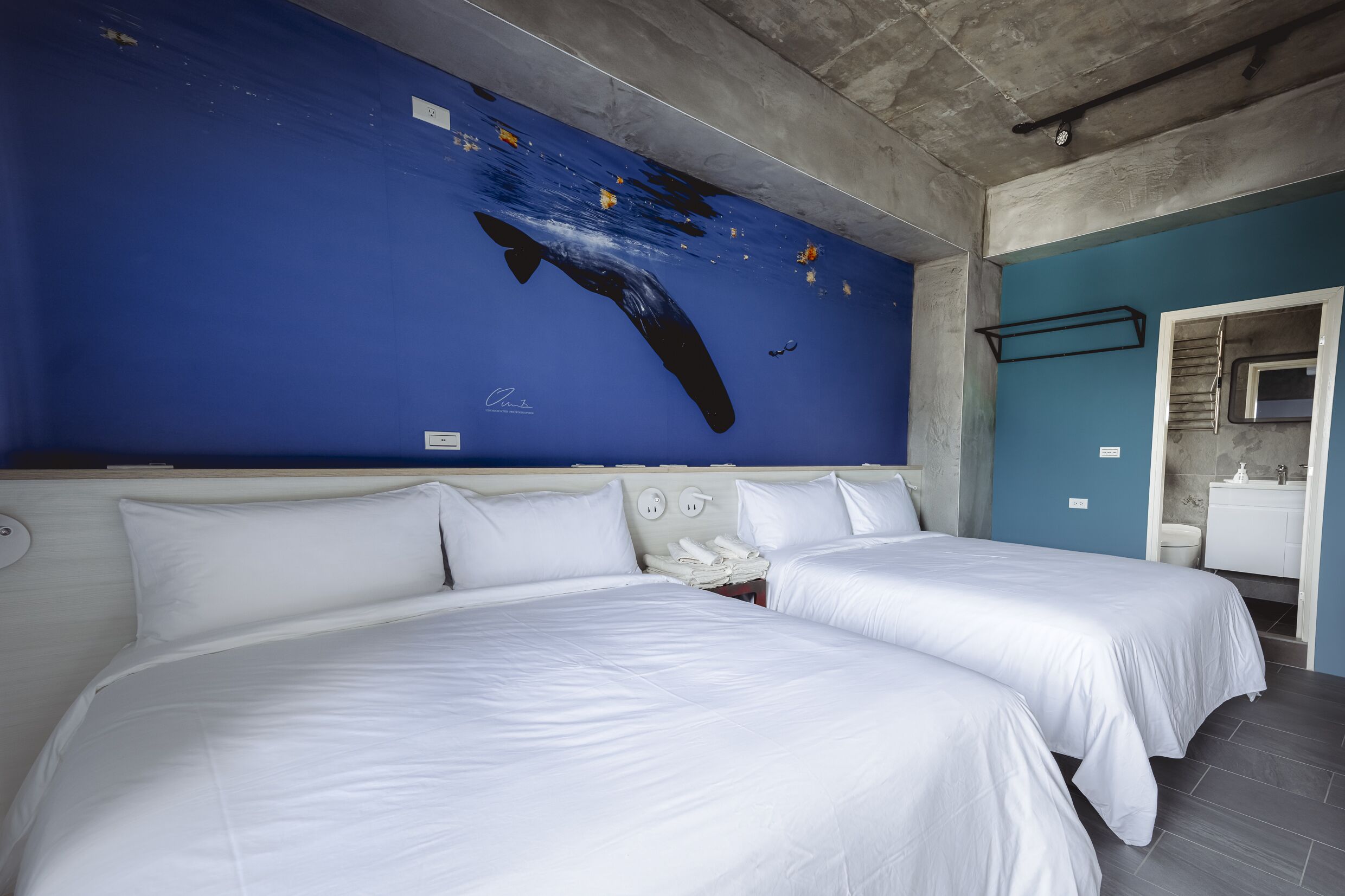 A hotel room with two beds, a blue wall mural, a white headboard, and a bathroom visible through an open door.