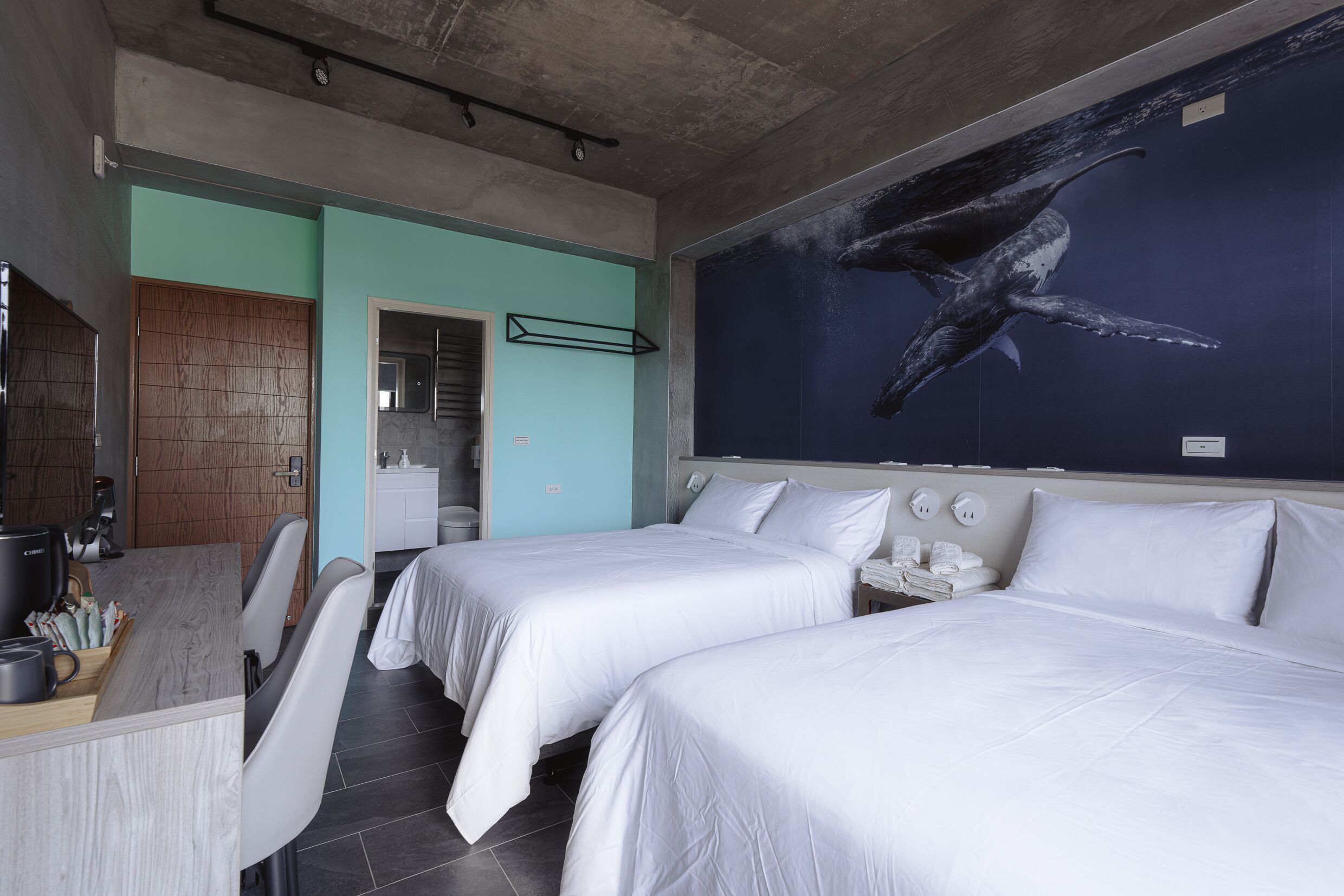 Habitación de hotel con dos camas, un escritorio con silla, un mural grande de una ballena y un baño visible a través de una puerta abierta.