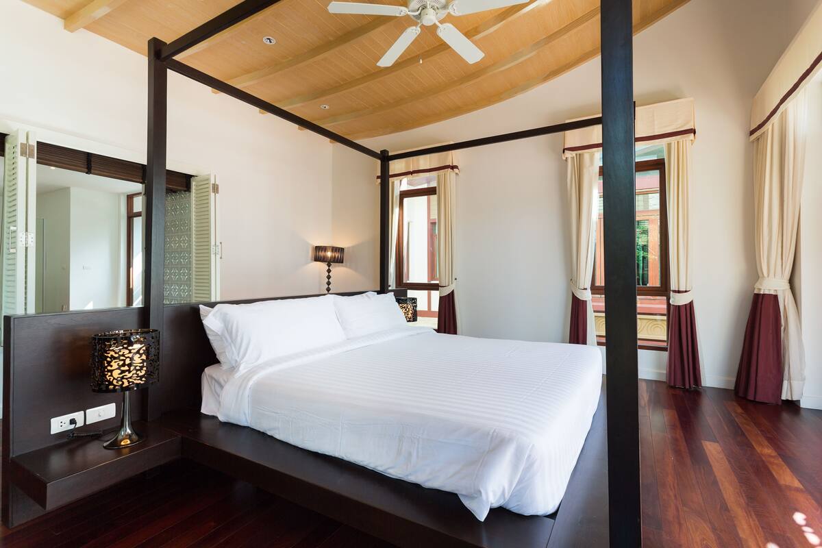 A four-poster bed with a wooden ceiling fan, a bedside table with a lamp, and a large mirror.