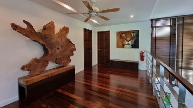 A spacious hallway with a wooden floor, a ceiling fan, a large wooden sculpture, and a painting of a lion on the wall. - Amatapura Beach Villa 12 (Krabi)