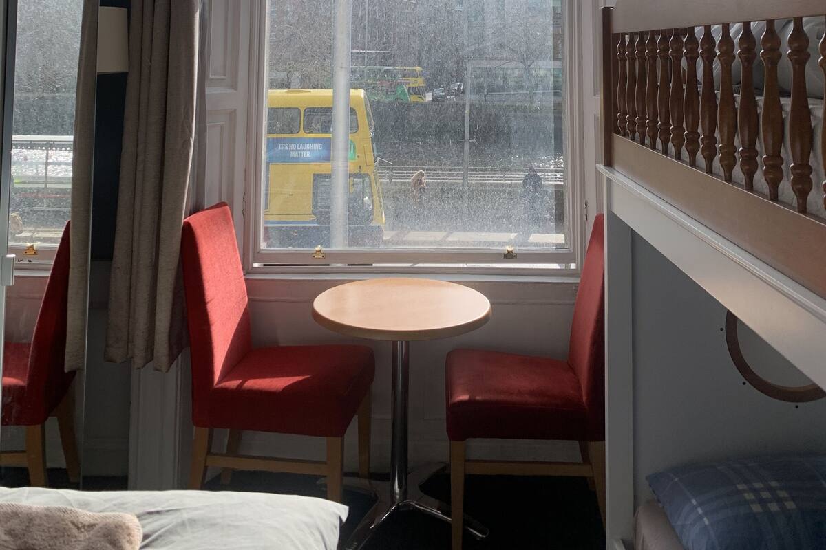 Une pièce avec une grande fenêtre, deux chaises rouges, une petite table ronde et un lit superposé avec une tête de lit en bois.
