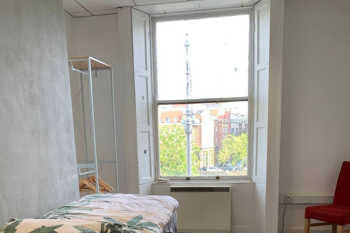 Une chambre à coucher avec un lit, un radiateur, une fenêtre donnant sur des immeubles et un fauteuil rouge.