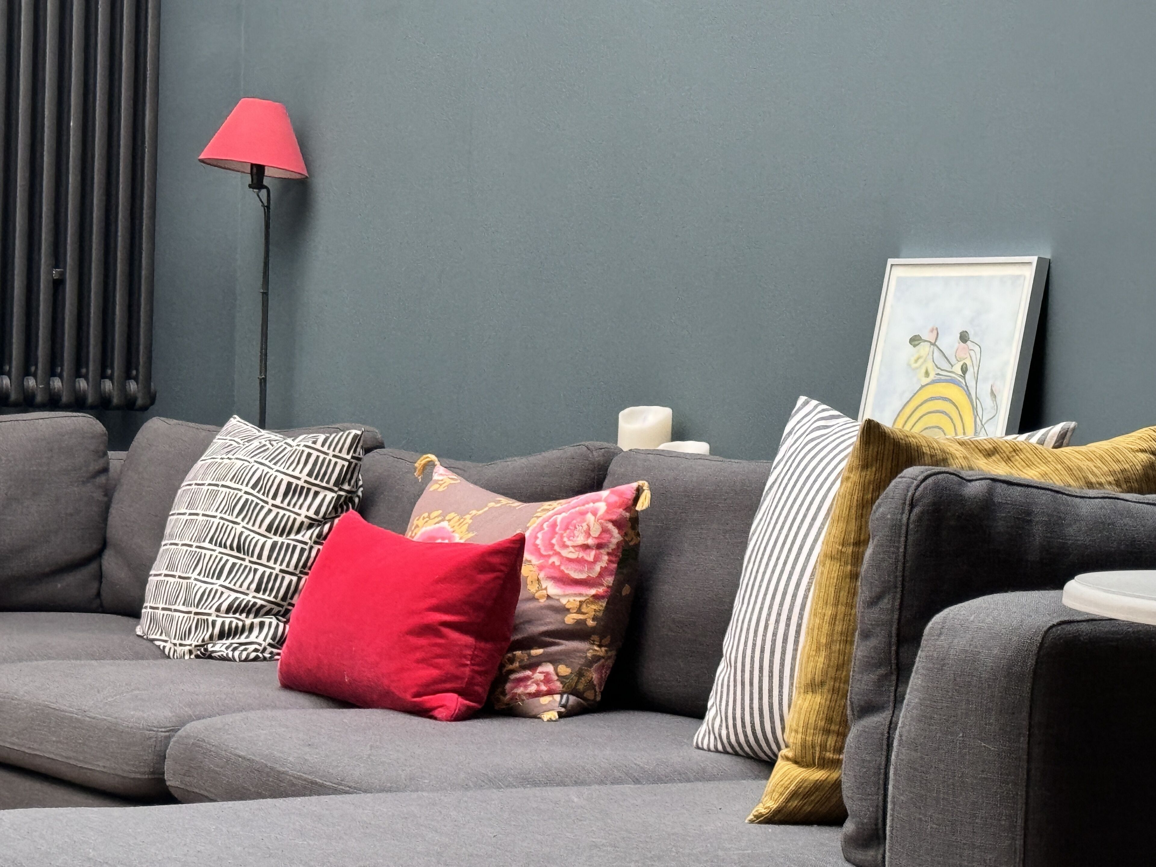 A grey sofa with various patterned cushions, a red lamp, and a framed artwork on the wall.