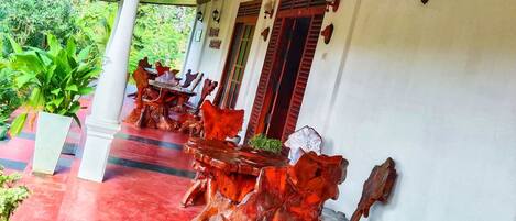 A covered outdoor area with a red floor, wooden tables with unique designs, and a white wall with windows and wall-mounted lights.